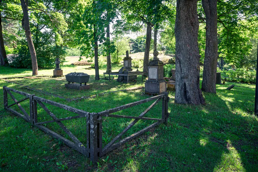 Historischer Friedhof mit Grabsteinen, umgeben von Bäumen und Grünflächen. Ein rustikales Zaunelement umrahmt den ruhigen Bereich.