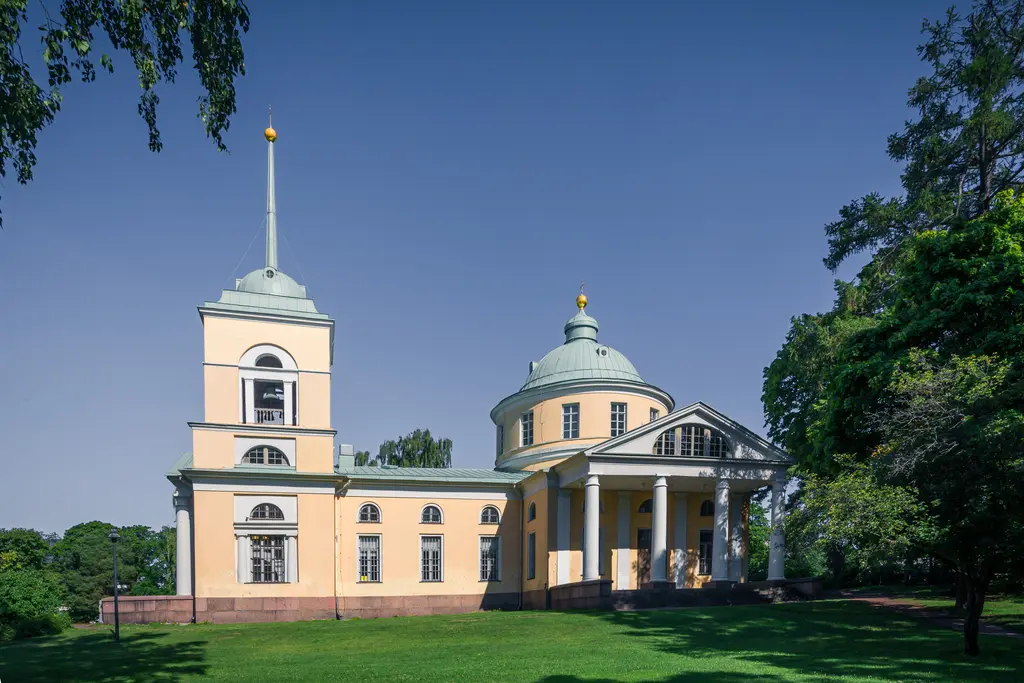 Stattliches Gebäude mit einer runden und einer rechteckigen Bauform, grünem Dach und goldenen Verzierungen, umgeben von Grünflächen.
