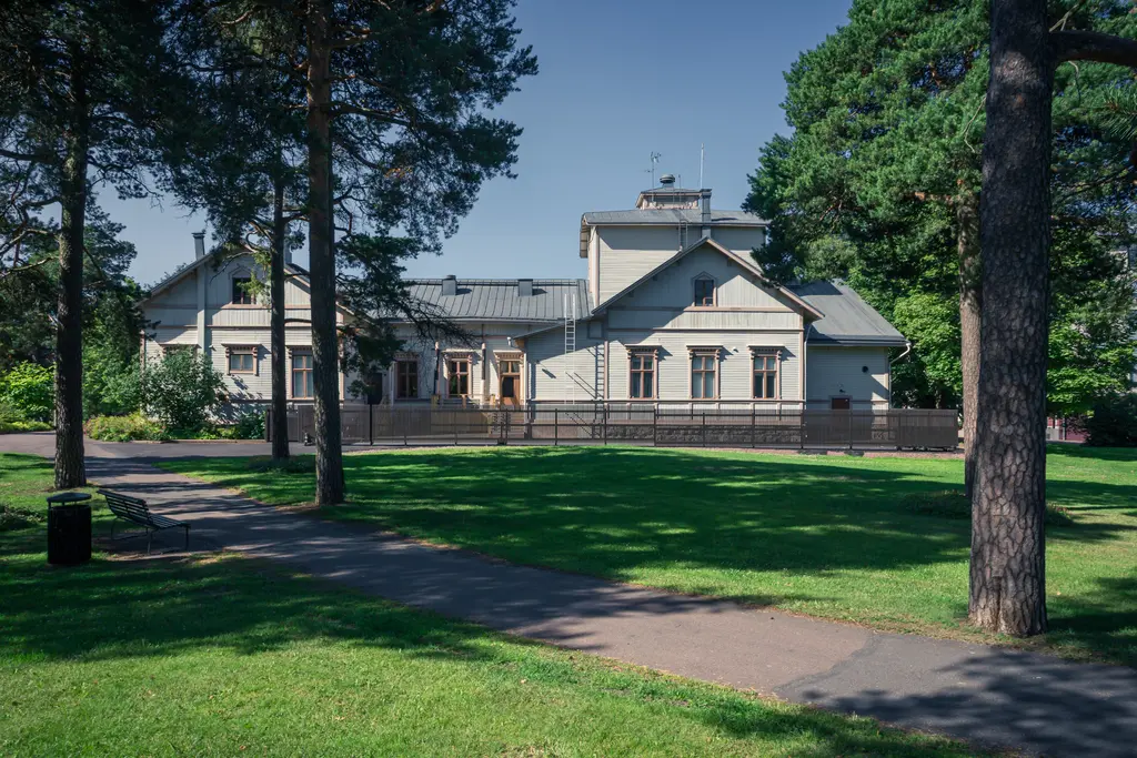 Historisches Holzgebäude mit einem Satteldach, umgeben von Bäumen und Rasenflächen, im Vordergrund ein Gehweg.