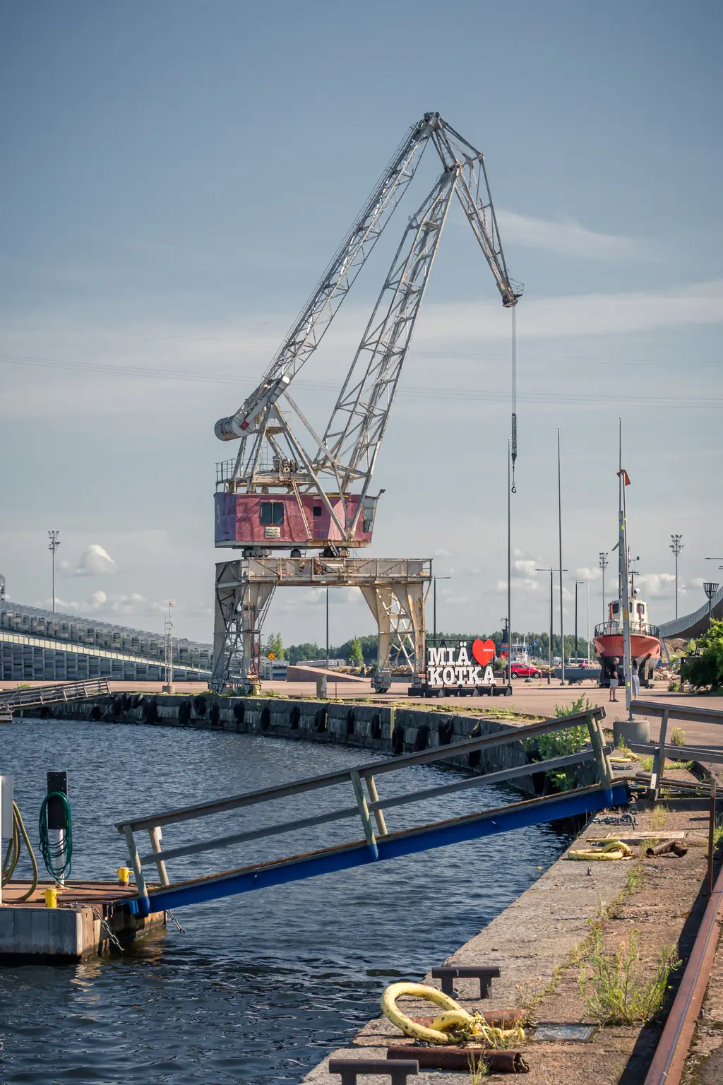 Hafenkran an einem schmalen Pier, im Hintergrund ein Schiff und eine bunte Liebeserklärung an Kotka. Ruhiges Wasser spiegelt die Szene.