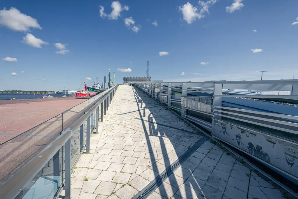Blick auf einen gepflasterten Weg, der zu einem erhöhten Aussichtspunkt führt. Links sind Boote und der Hafen sichtbar, dazu ein klarer blauer Himmel mit wenigen Wolken.