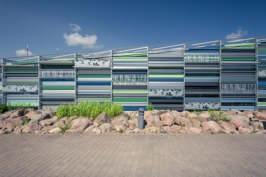 Futuristische Gebäude-Fassade mit bunten vertikalen Rechtecken in Blau, Grün und Grau, umgeben von Steinen und Gras.