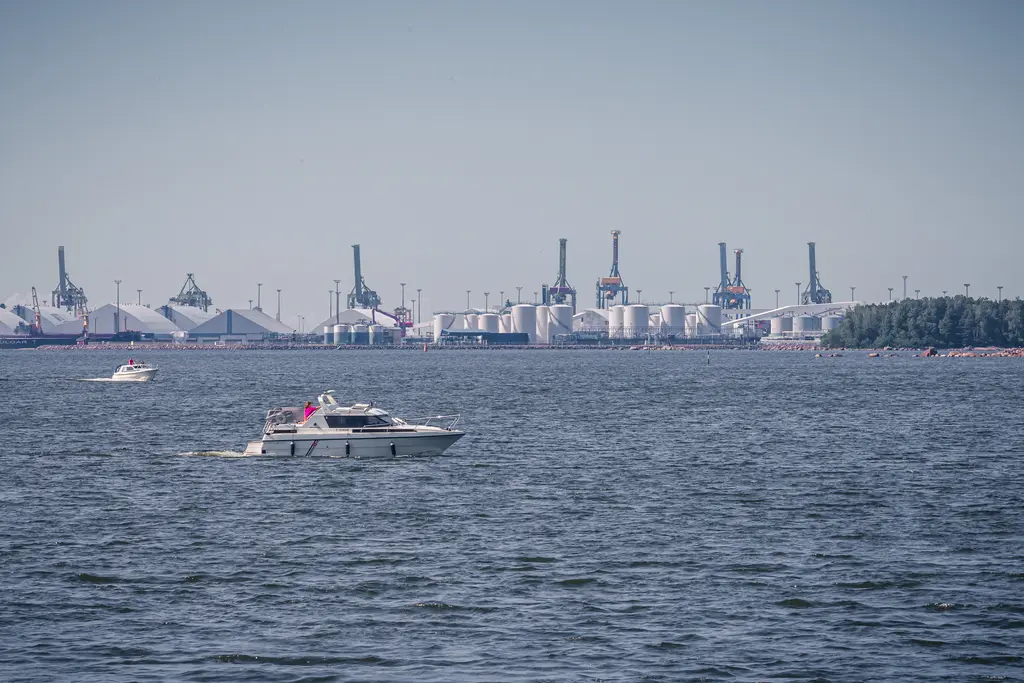 Boot auf dem Wasser mit Hafenkränen und Lagerhäusern im Hintergrund. Der Himmel ist klar und die Wasseroberfläche glitzert.