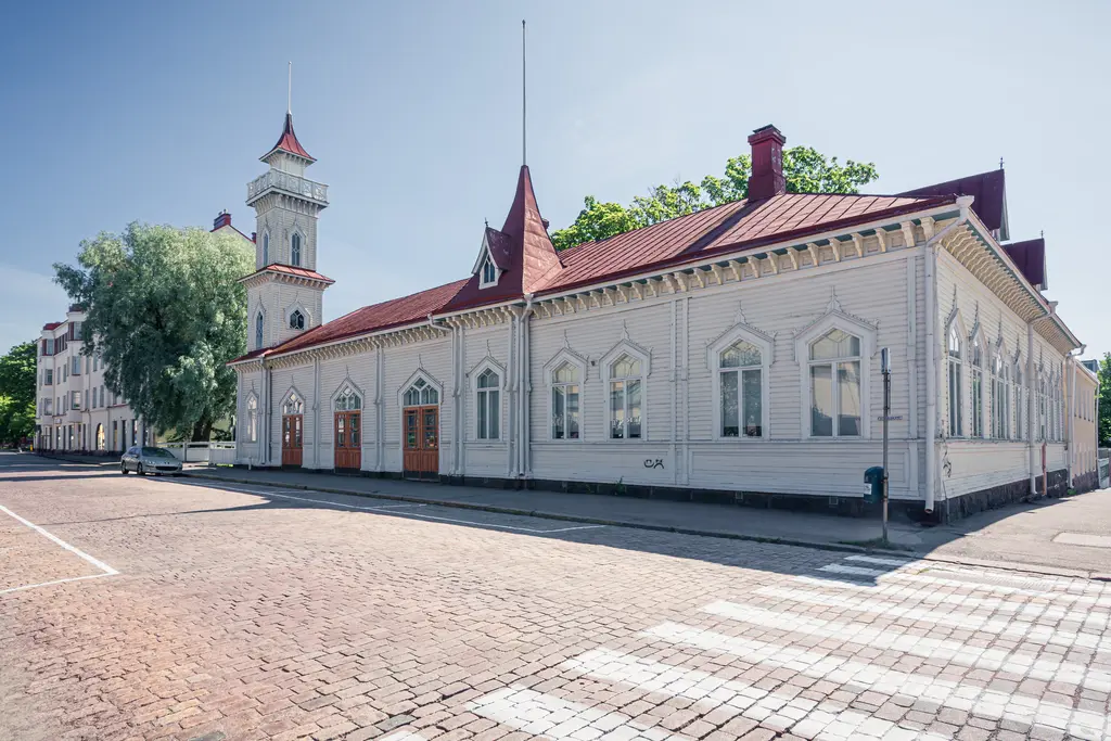 Historisches Holzgebäude mit rotem Satteldach und einem kleinen Uhrturm, umgeben von Bäumen und gepflasterter Straße.