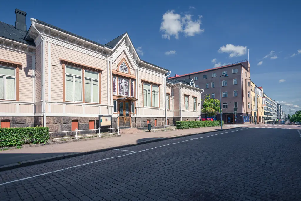 Historisches Gebäude mit dekorativen Elementen und großen Fenstern, umgeben von einer gepflasterten Straße und modernen Gebäuden.