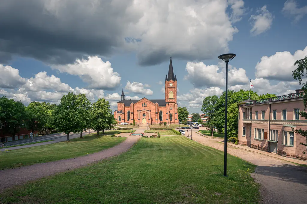 Kirche mit roten Ziegeln und hohen, spitzen Türmen, umgeben von Bäumen und einem grünen Rasen, unter einem bewölkten Himmel.