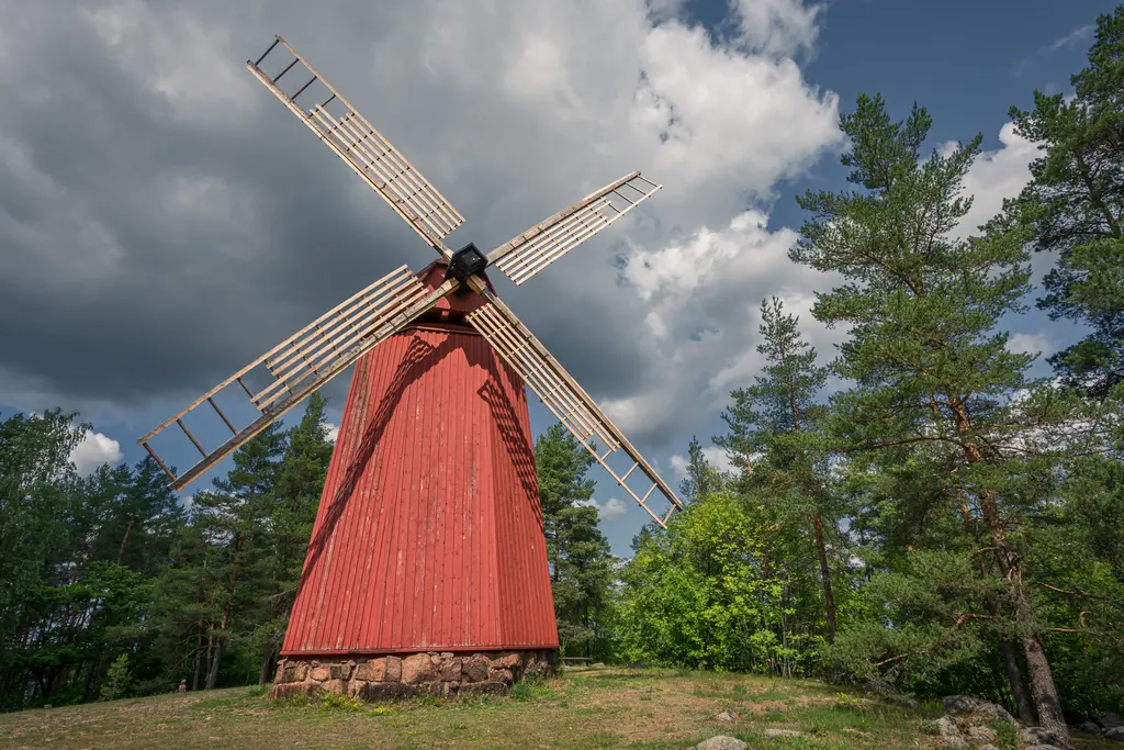 Roten Windmühle mit großen, beweglichen Flügeln steht auf einer Wiese, umgeben von grünen Bäumen und einem bewölkten Himmel.