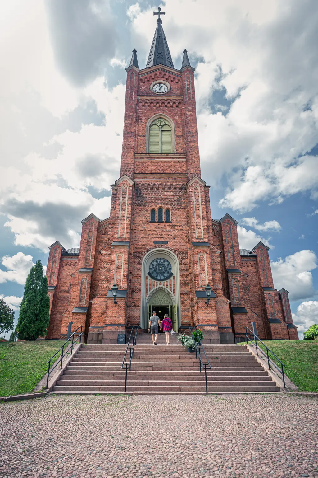 Historische Kirche aus roten Ziegeln mit hohem Turm, umgeben von Stufen, auf denen zwei Personen zur Eingangstür gehen.