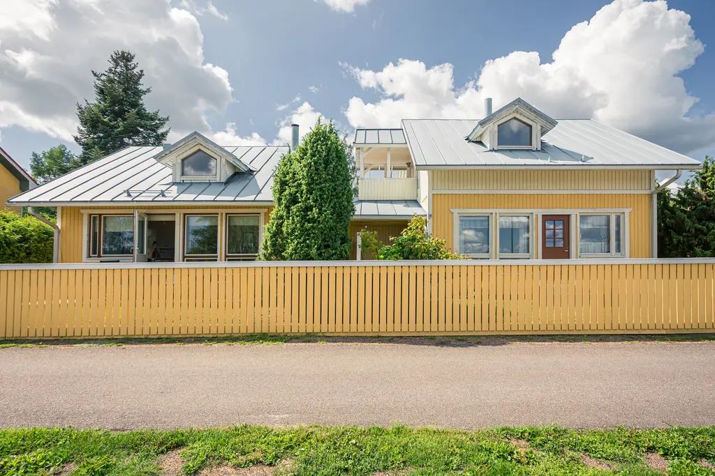 Gelbes Holzhaus in Loviisa mit einem grünen Garten, umgeben von einer weißen Holzhecke. Der Himmel ist mit einigen Wolken versehen.
