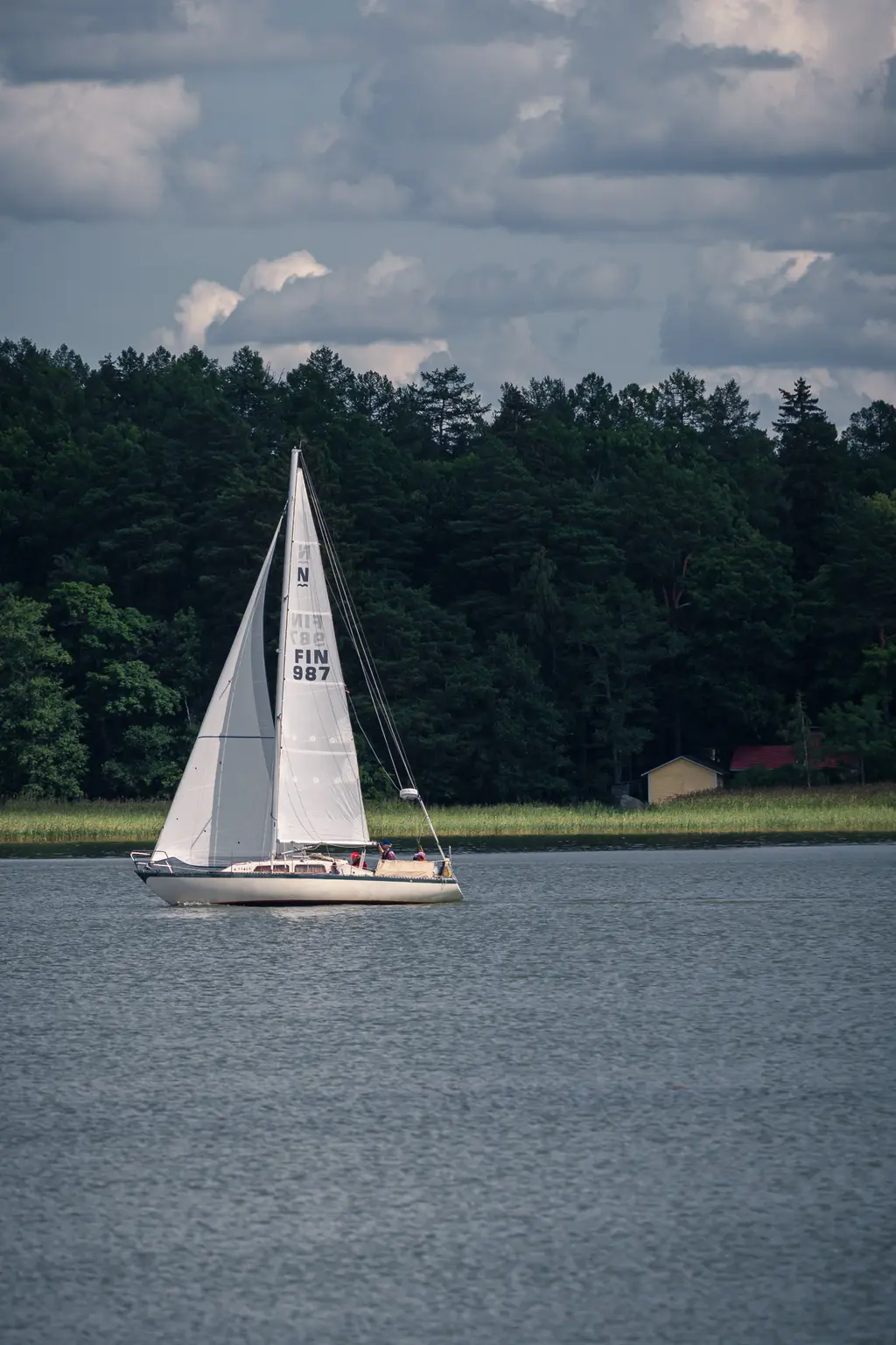 Segelboot auf ruhigem Wasser bei Loviisa, umgeben von grünen Bäumen und einem kleinen gelben Haus im Hintergrund.