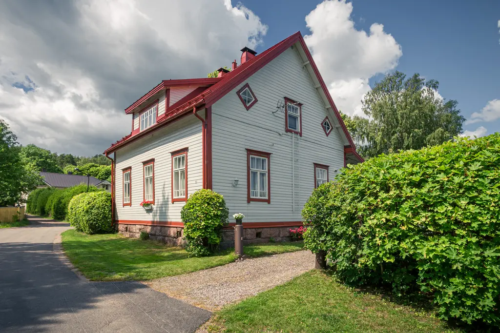 Helles, zweigeschossiges Holzhaus in Loviisa mit rotem Dach und Fensterrahmungen. Umgeben von grünen Sträuchern und einem Gehweg.