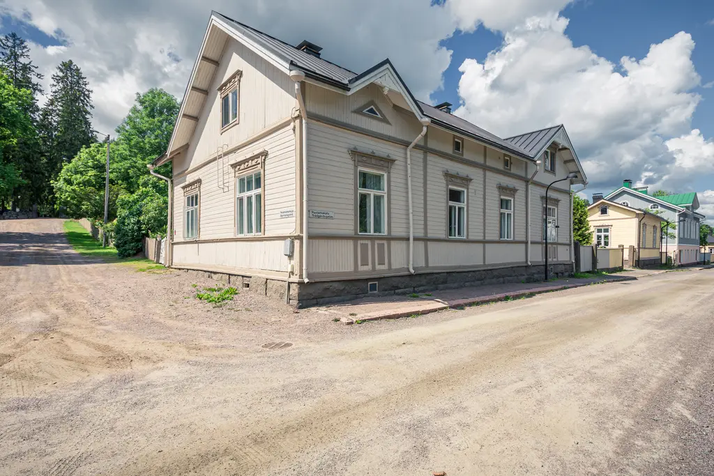 Historisches Holzhaus in Loviisa mit weißen Wänden und schmalen Fenstern, umgeben von Bäumen und einer kiesbeladenen Straße.