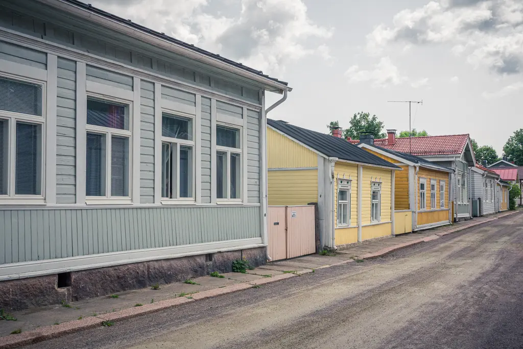 Historische Holzgebäude an einer stillen Straße, mit grauen und gelben Fassaden sowie typischen Fenstern. Der Gehweg ist uneben.