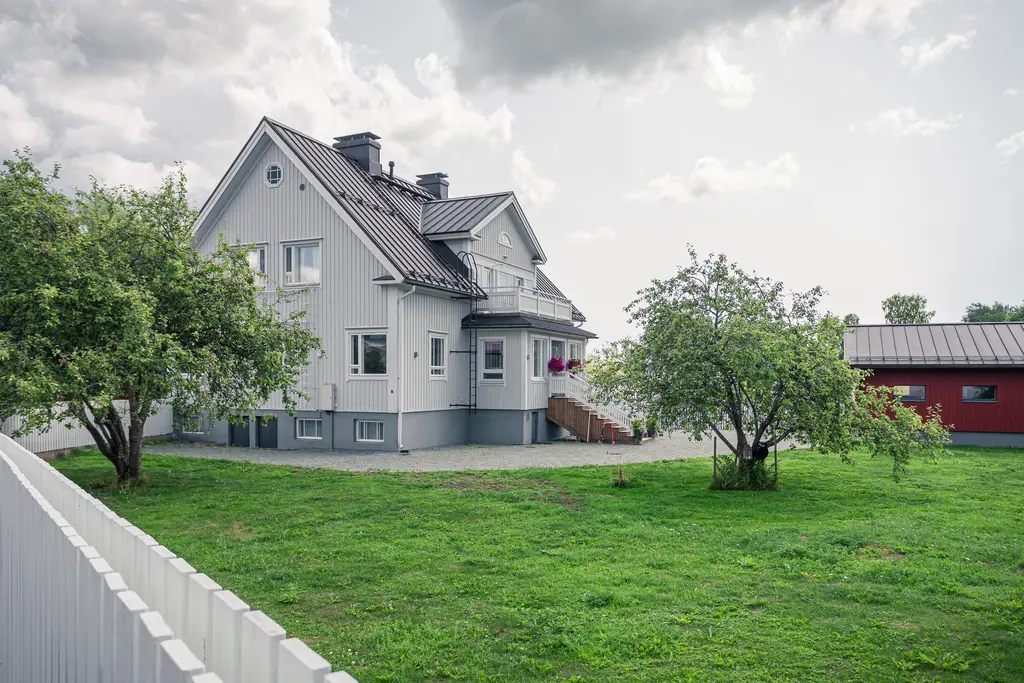 Helles Holzhaus mit grauer Fassade und einem Satteldach, umgeben von Rasen und einigen Bäumen. Im Hintergrund steht ein rotes Nebengebäude.