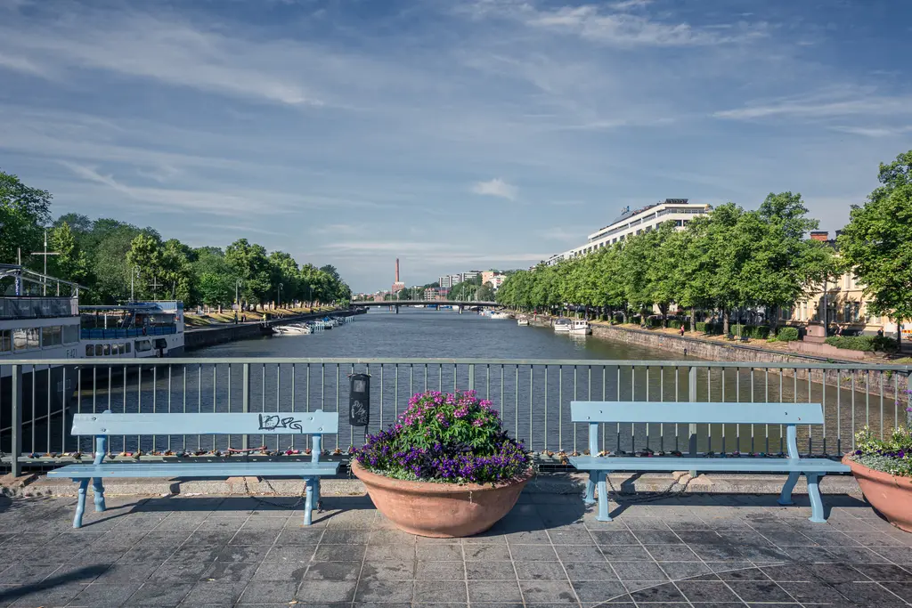 Blick auf einen ruhigen Fluss mit Bänken am Ufer, umgeben von Bäumen und Gebäuden. Ein Blumenbehälter steht im Vordergrund.