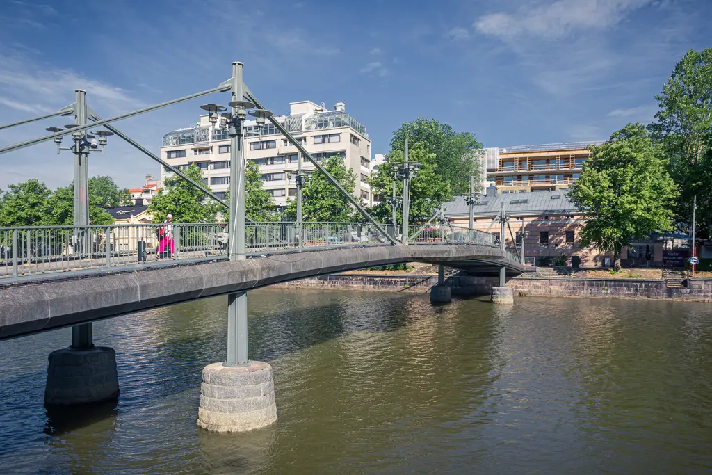 Eisenbahnbrücke über einen ruhigen Fluss mit angrenzenden Bäumen und modernen Gebäuden im Hintergrund. Ein Fußgänger ist auf der Brücke zu sehen.