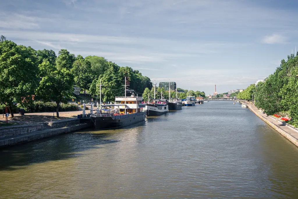 Hafenansicht mit mehreren Booten am Ufer, umgeben von grünen Bäumen. Ein ruhiges Wasser fließt durch die Szene unter blauem Himmel.