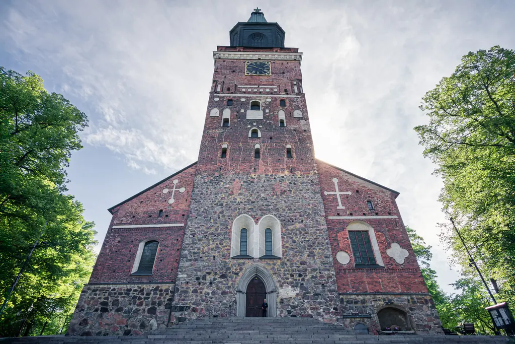 Historische Kirche aus roten Ziegeln mit markantem Turm, umgeben von grünen Bäumen und einer hellen Himmelshintergrund.