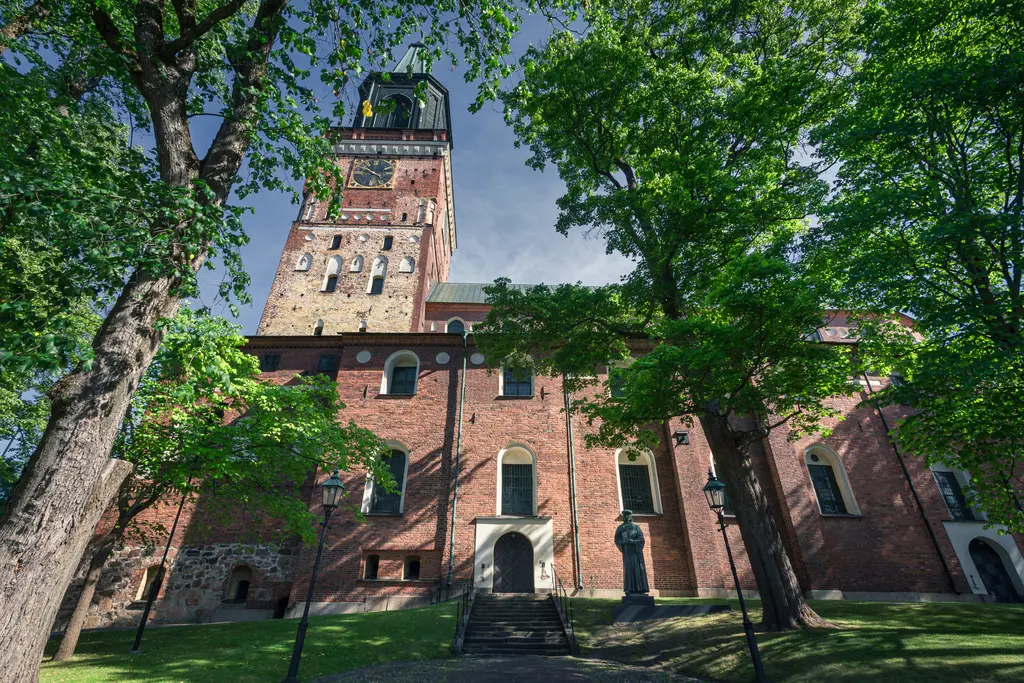 Historisches Backsteingebäude mit einem hohen Turm, umgeben von grünen Bäumen. Stufen führen zur Eingangstür.