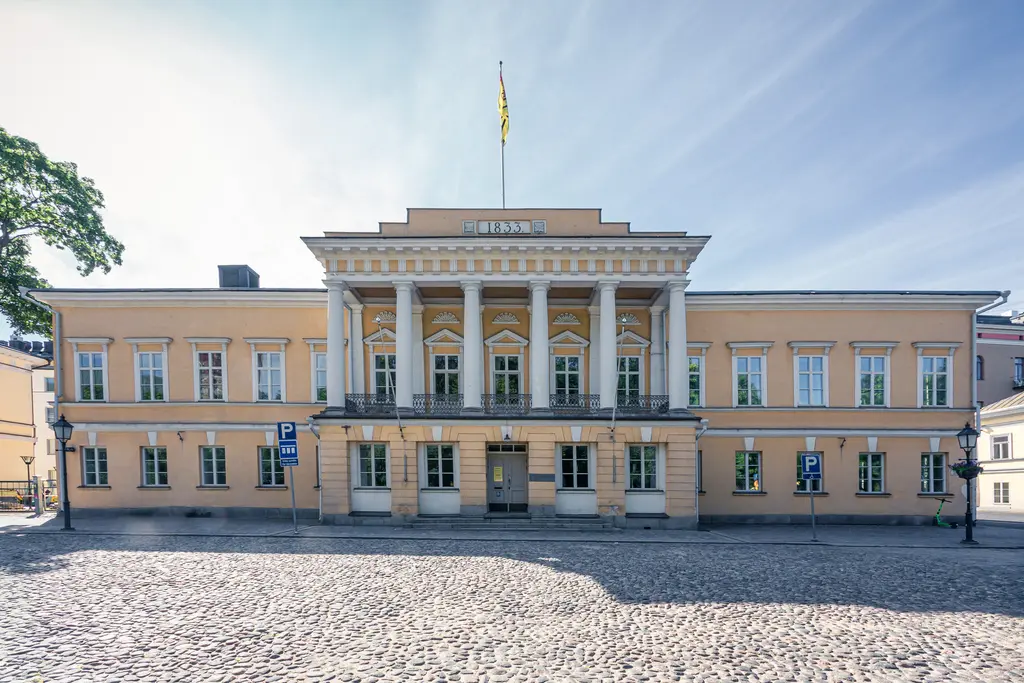 Historisches Gebäude mit Säulenfassade und orange gefärbter Wand. Vor dem Gebäude verläuft ein gepflasterter Platz mit Parkplätzen.