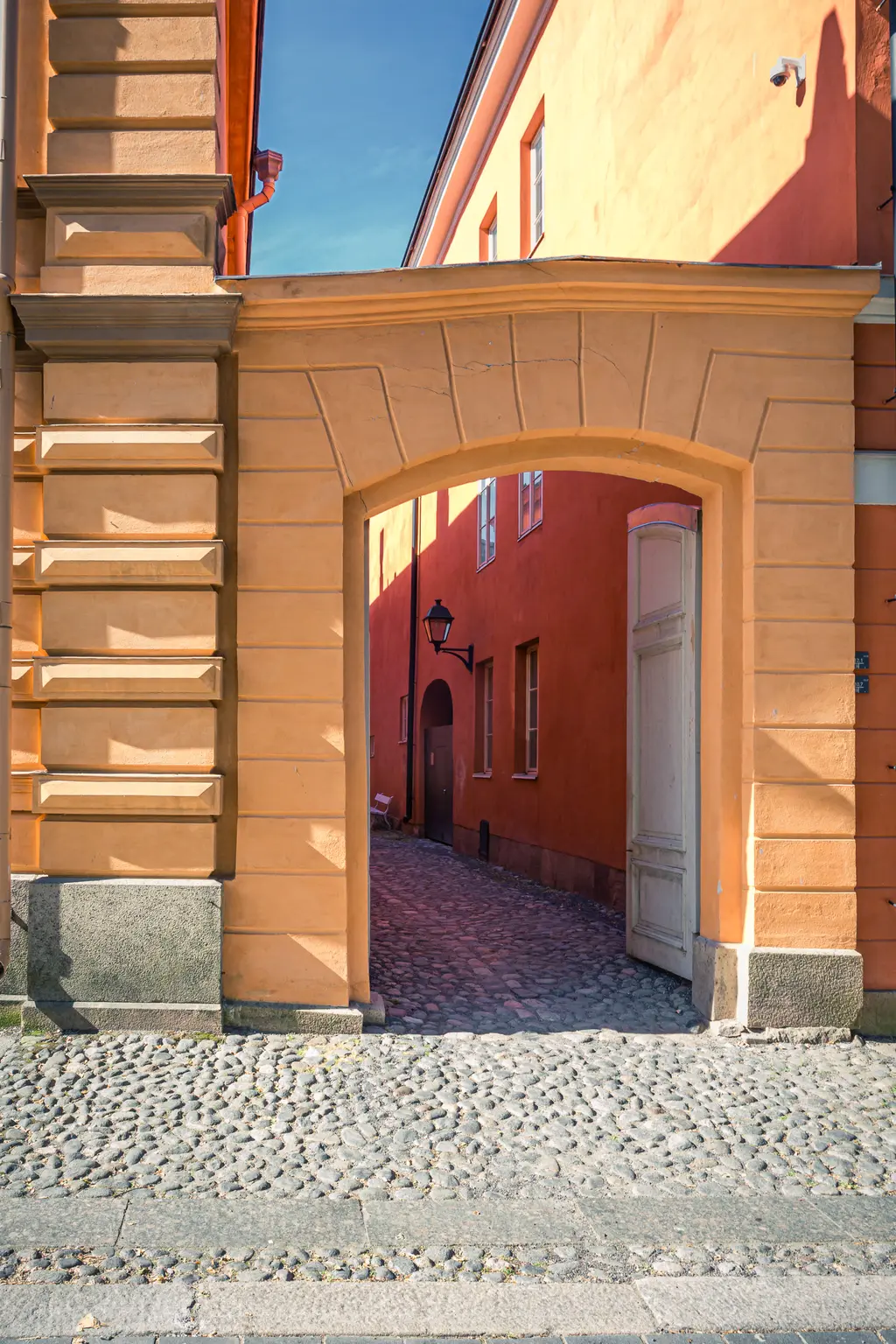 Durch einen hohen, bogenförmigen Eingang ist eine enge Gasse mit gepflastertem Boden sichtbar. Die Wände der umgebenden Gebäude sind in warmen Orange- und Gelbtönen gehalten. Eine Laterne hängt an der Wand.