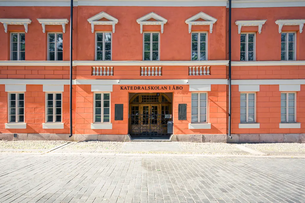 Rosa Gebäude mit weißem Rahmen, fünf Fenstern und einer Eingangstür in der Mitte. Über der Tür steht „Katedralskolan Åbo“.