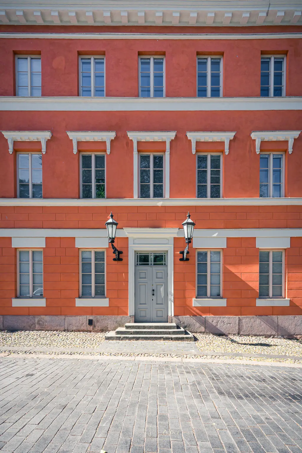 Fassade eines Hauses in warmem Orange mit zwei Lampen neben dem Eingang. Das Gebäude hat mehrere Fenster und eine graue Tür.