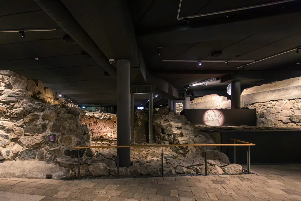 Archäologische Ausgrabungen in einem modernen Innenraum, mit sichtbaren Steinen und Ruinen, unter Beleuchtung und einer Holzbrüstung.