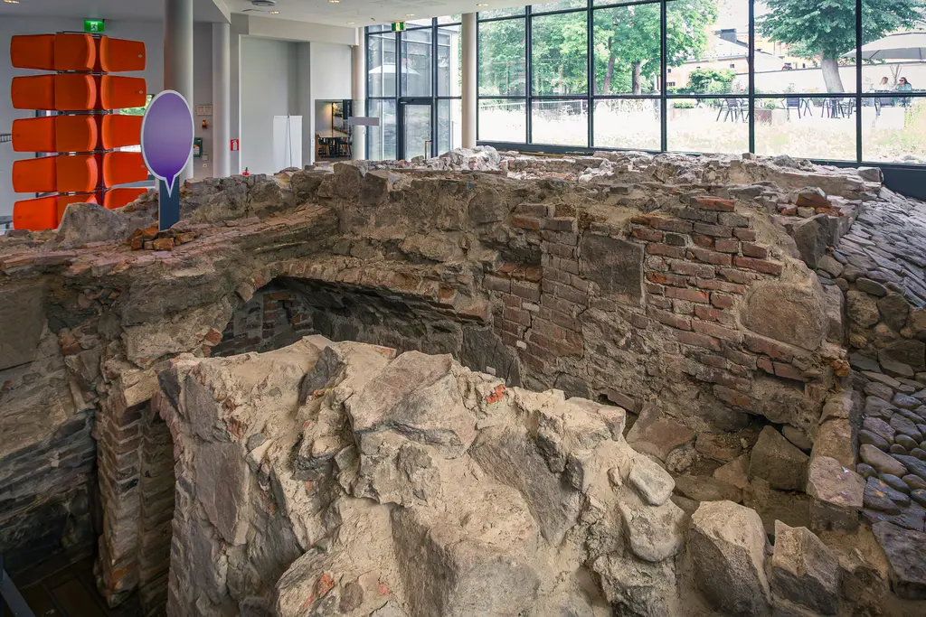 Ruinen einer alten Mauer aus Stein und Ziegel, teilweise unvollständig. Im Hintergrund große Fenster und moderne Architektur mit natürlichem Licht.
