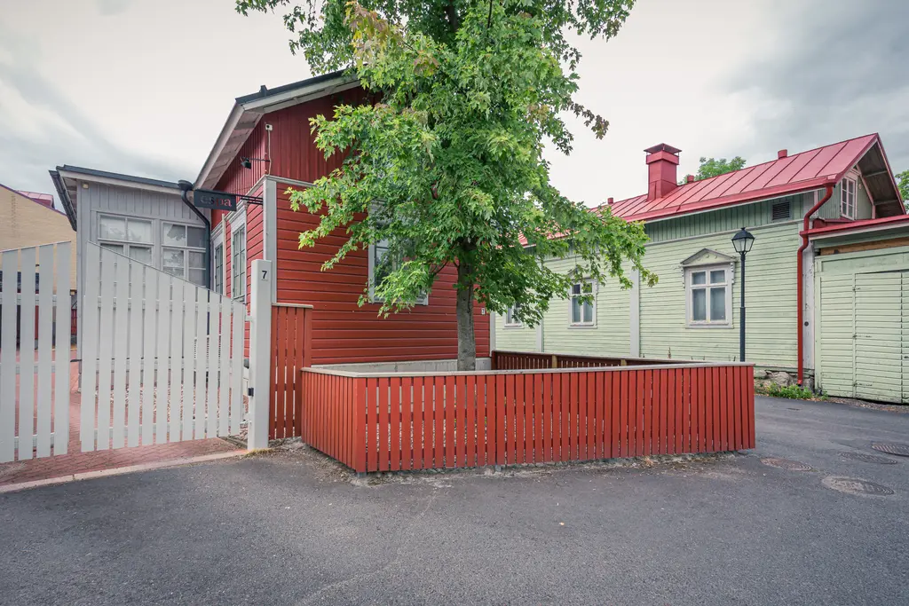 Rote holzverkleidete Häuser, umgeben von einem kleinen Garten mit einem Baum und einem weißen Zaun, auf einer ruhigen Straße.