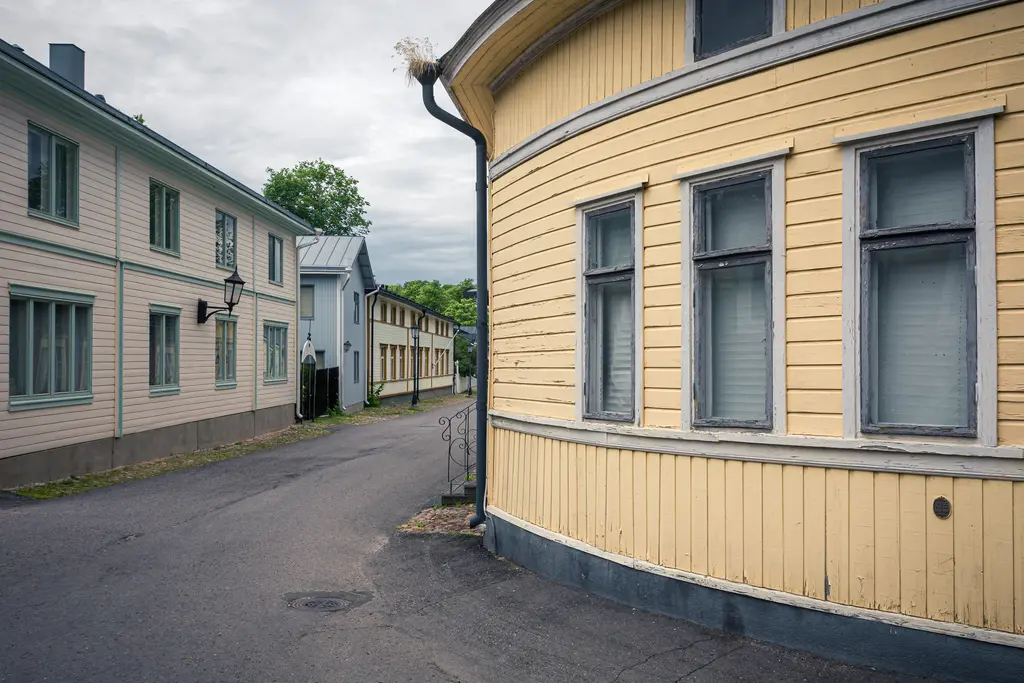 Historische, bunt gestrichene Holzhäuser in einer engen Straße, die um eine Kurve verläuft. Die Fassaden zeigen Fenster mit grauen Rahmen.
