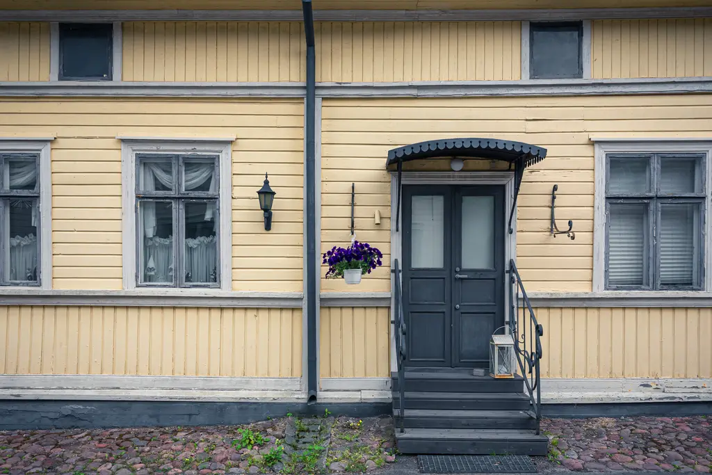 Gelbe Holzwand mit schwarzen Fenstern und einer dunklen Eingangstür. Vor der Tür hängt ein Blumenkasten mit lila Blumen.