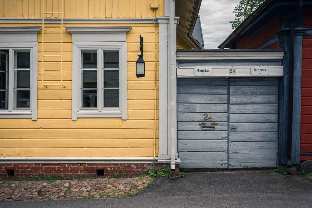 Gelbe Holzfassade mit zwei Fenstern und einer Laterne, daneben eine graue Tür mit Nummern. Steine und Gras am Boden.