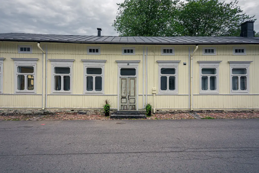 Gelbes Holzgebäude mit mehreren Fenstern und einer zentralen Eingangstür, umgeben von einem gepflasterten Weg und bepflanzten Kübeln.