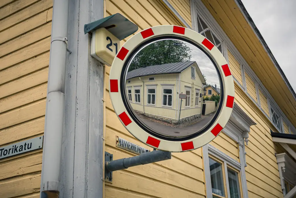 Verkehrsspiegel an einer gelben Holzfassade, der ein gegenüberliegendes Gebäude und die Straße spiegelt.