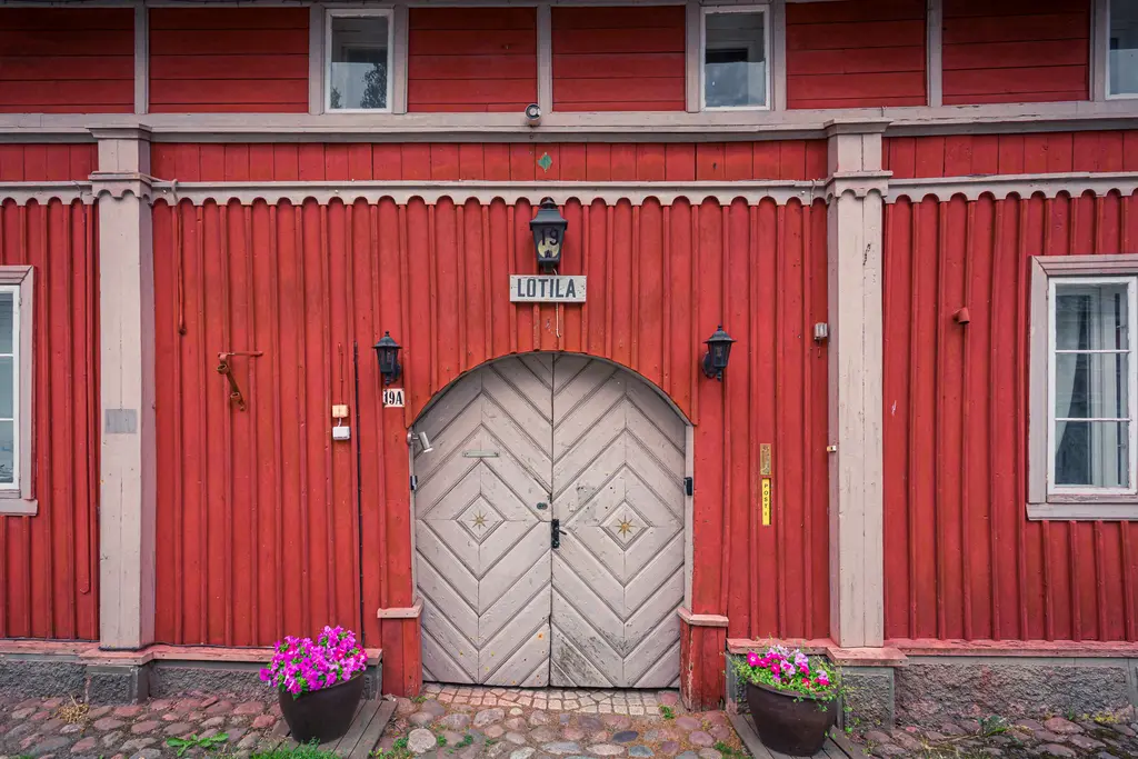 Rote Holzfassade mit großer, gewölbter Doppeltür, die mit einem Ornament verziert ist. Über der Tür prangt ein Schild mit der Aufschrift „LOTILA“. Blumenkübel mit bunten Blumen stehen neben der Tür.