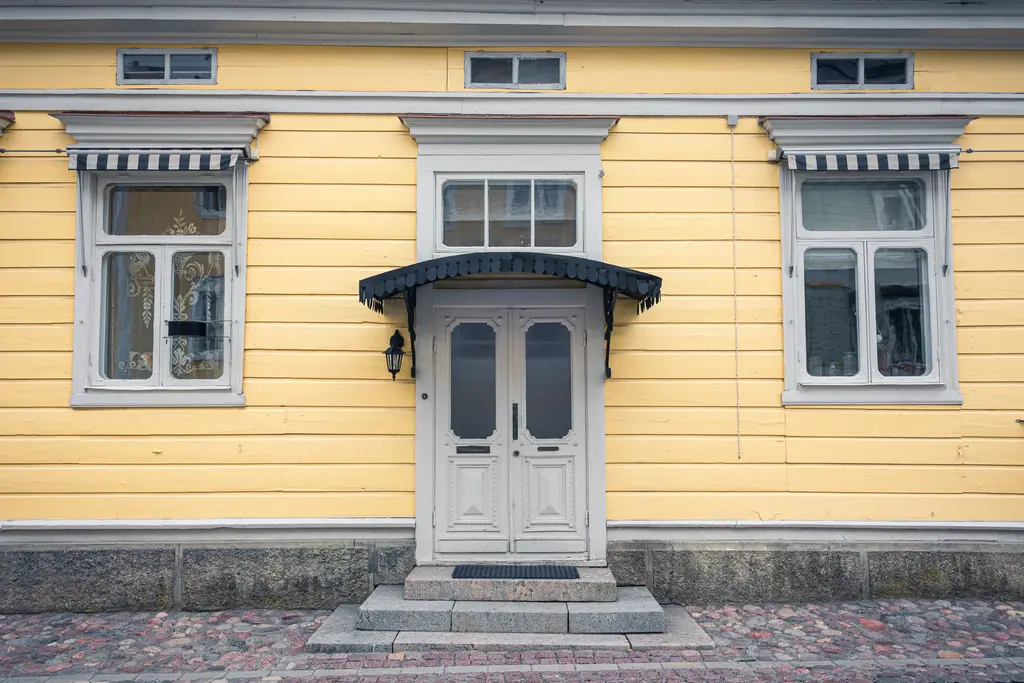 Gelbe Holzfassade mit einer weißen Doppeltür und einem Vordach. Links und rechts sind Fenster mit weißen Rahmen und gestreiften Jalousien.