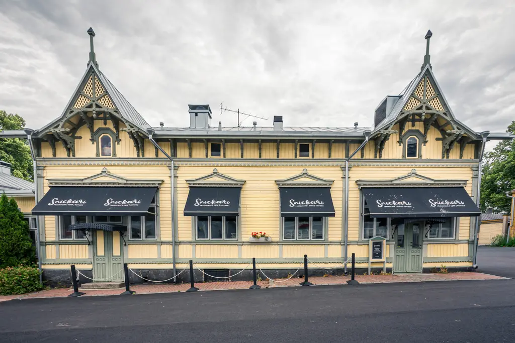 Historisches, gelbes Holzgebäude mit aufwendigen architektonischen Details, zwei Eingängen und schwarzem Sonnenschutz.
