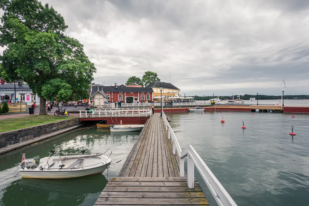 Holzsteg am Hafen mit kleinen Booten und umgebenden Gebäuden. Grüne Bäume und ein bewölkter Himmel sind im Hintergrund sichtbar.