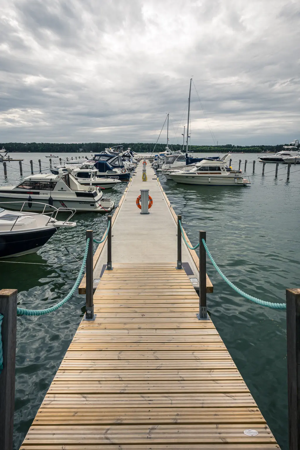 Steg am Hafen mit mehreren Booten, die am Wasser festgemacht sind. Der Steg ist aus Holz und hat grüne Seile an den Seiten. Der Himmel ist bewölkt.