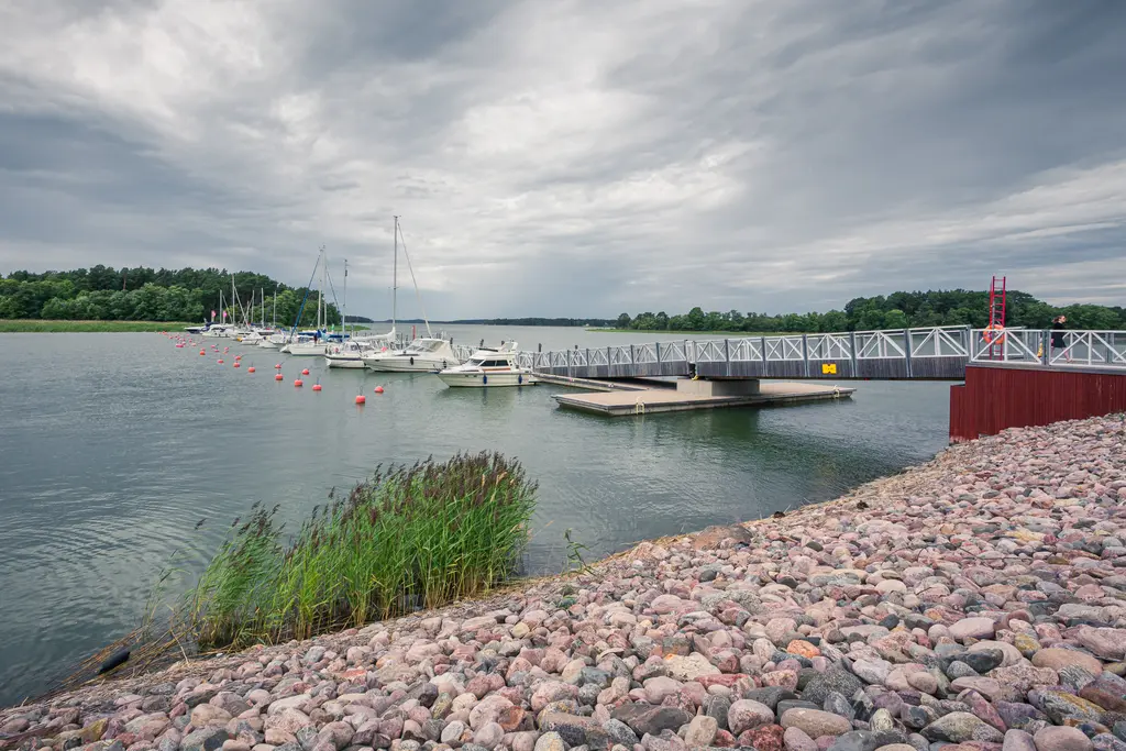 Hafenansicht mit einem Steg, an dem mehrere Segelboote festgemacht sind. Im Vordergrund eine Uferböschung mit Kies und Schilf.