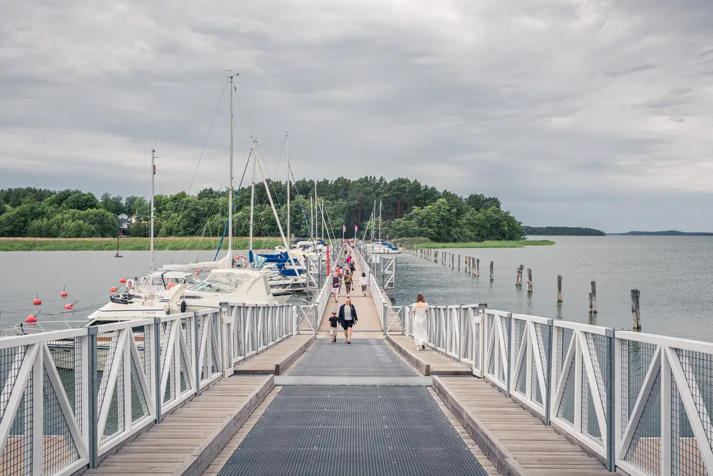 Pier mit Booten, umgeben von Wasser und Bäumen. Menschen flanieren auf dem Holzsteg, der ins Wasser führt. Wolkiger Himmel.