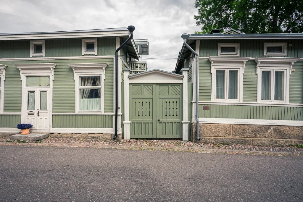 Zwei grüne Holzhäuser mit weißen Fensterrahmen und Türen stehen einander gegenüber. Zwischen ihnen befindet sich ein geschlossener Holztor.