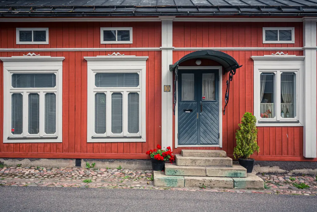 Rotes Holzhaus mit weißem Fachwerk, blauen Fenstern und einer grauen Eingangstür. Blühende Pflanzen stehen am Eingang.