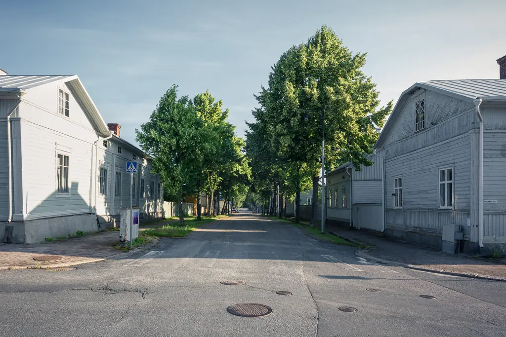 Schmale Straße zwischen alten, hellen Holzhäusern, flankiert von grünen Bäumen. Sanfte Lichtverhältnisse, ruhige Atmosphäre.