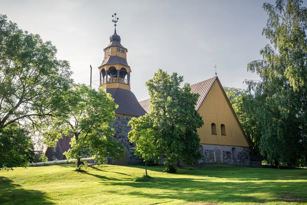 Historische Kirche mit auffälligem Turm, umgeben von grünen Bäumen und Wiese. Der Turm hat eine Holzverkleidung und dekorative Elemente.