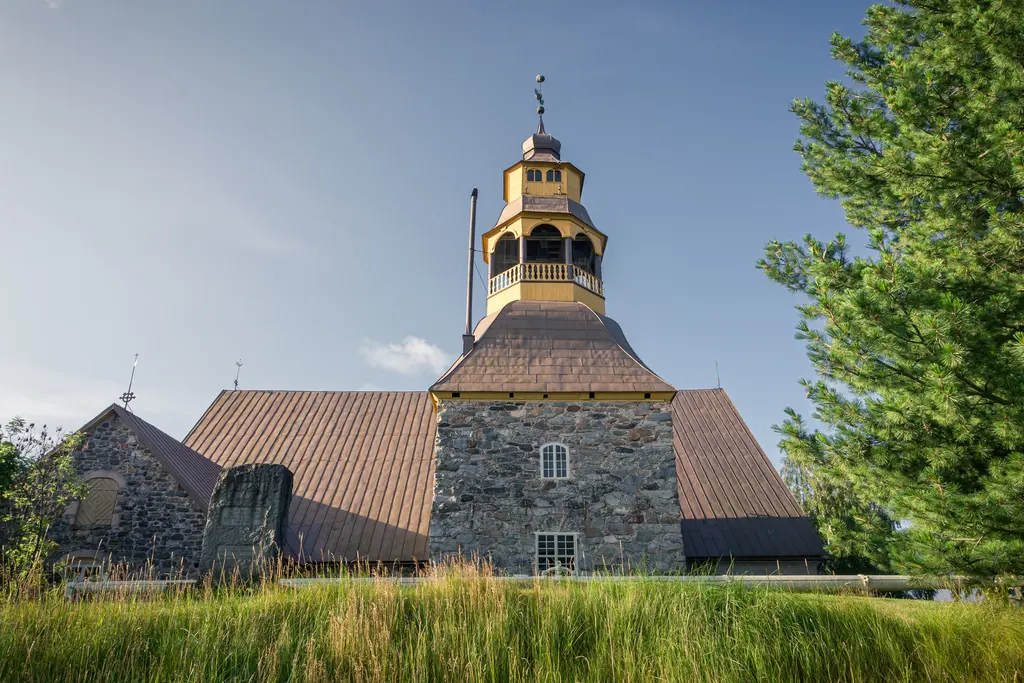 Historisches Gebäude mit steilem Dach und einem markanten Turm. Umgeben von grünem Gras und Bäumen, unter blauem Himmel.