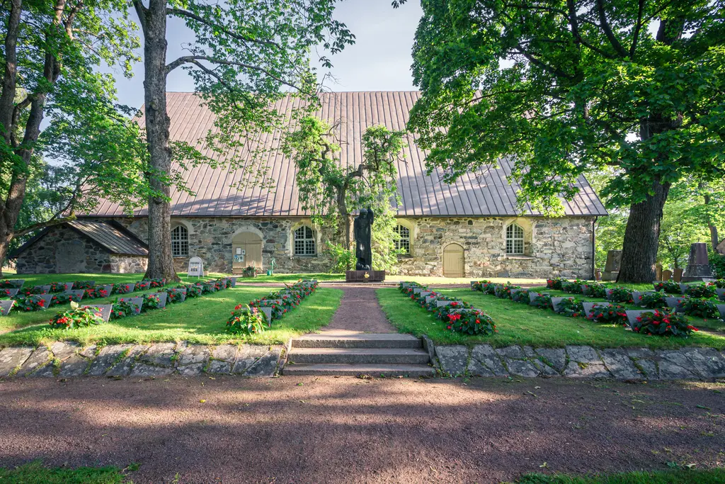 Steinernes Gebäude mit einem Satteldach, umgeben von grünen Bäumen und bunten Blumenbeeten. Ein gepflegter Weg führt zur Eingangstür.