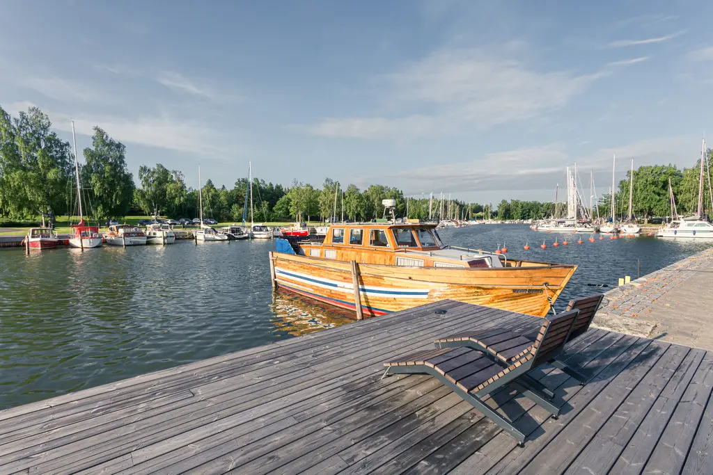 Holzboot am Steg, umgeben von Booten im Hafen. Im Hintergrund sind Bäume und ein blauer Himmel sichtbar. Eine Liege ist am Steg platziert.