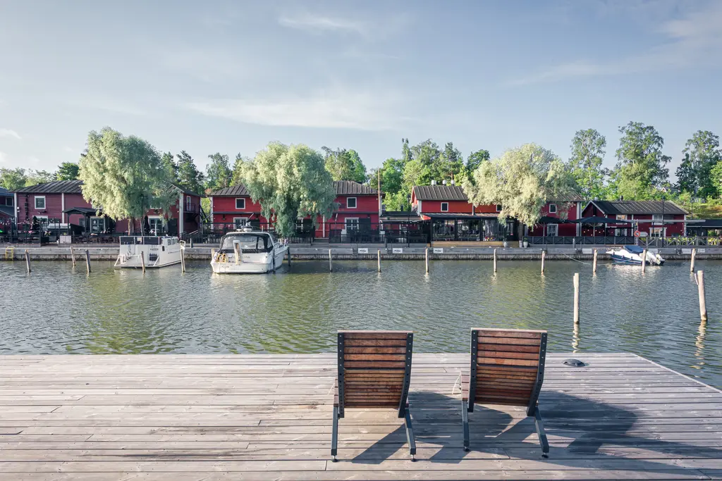 Zwei Holzliegestühle auf einem Steg mit Blick auf ein ruhiges Gewässer, umgeben von roten Häusern und grünen Bäumen.